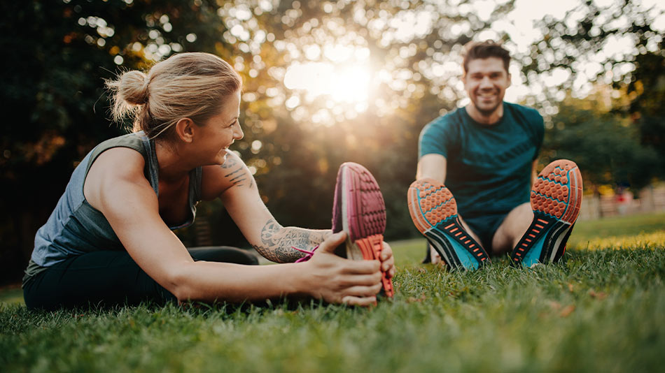 Blonde vrouw en brunette man strekken hun benen na het hardlopen. Zittend op de grasvloer, glimlachend.